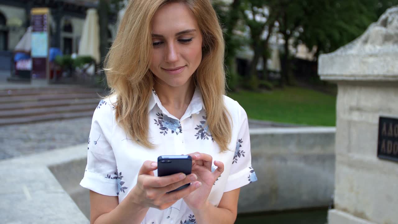 Young girl using smartphone sitting on fountain in the city. Happy young woman smiling and using iPhone smart phone camera. Tourism, instagram, modern technology.
