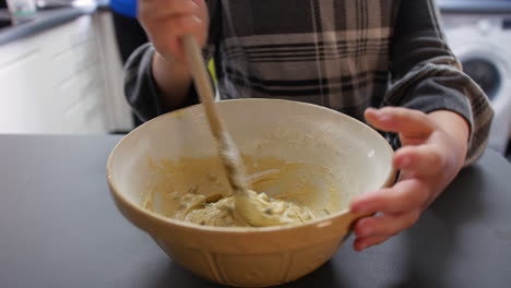 a girl mixing cookie dough cake mixture in a bowl at home in a kitchen with a wooden spoon