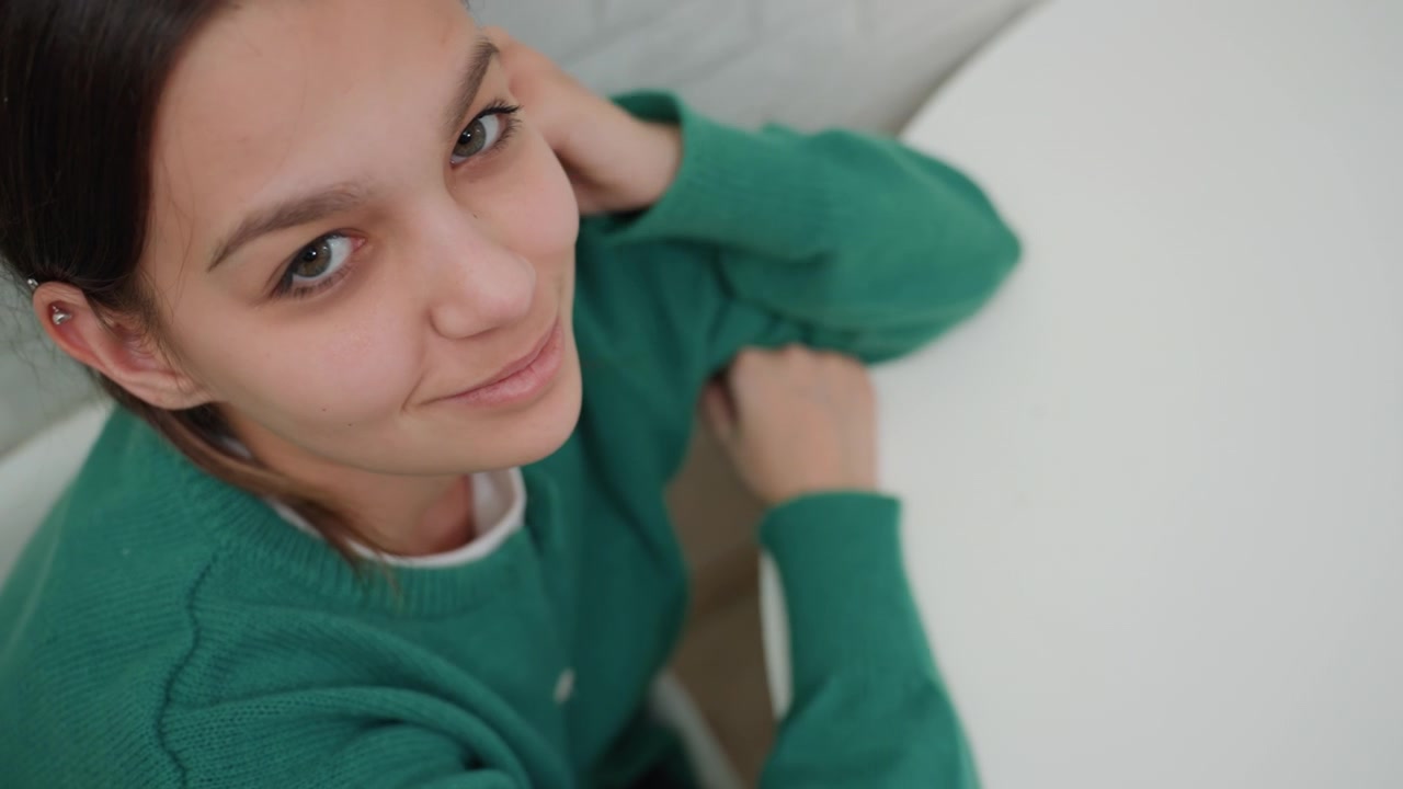 Giovane donna al tavolo, nuovo appartamento. Studentessa giocosa si ferma durante il giorno del trasloco, appoggiata al tavolo e facendo gesti espressivi con le mani mentre chiacchiera, indossando un comodo maglione verde, luce naturale soffusa.