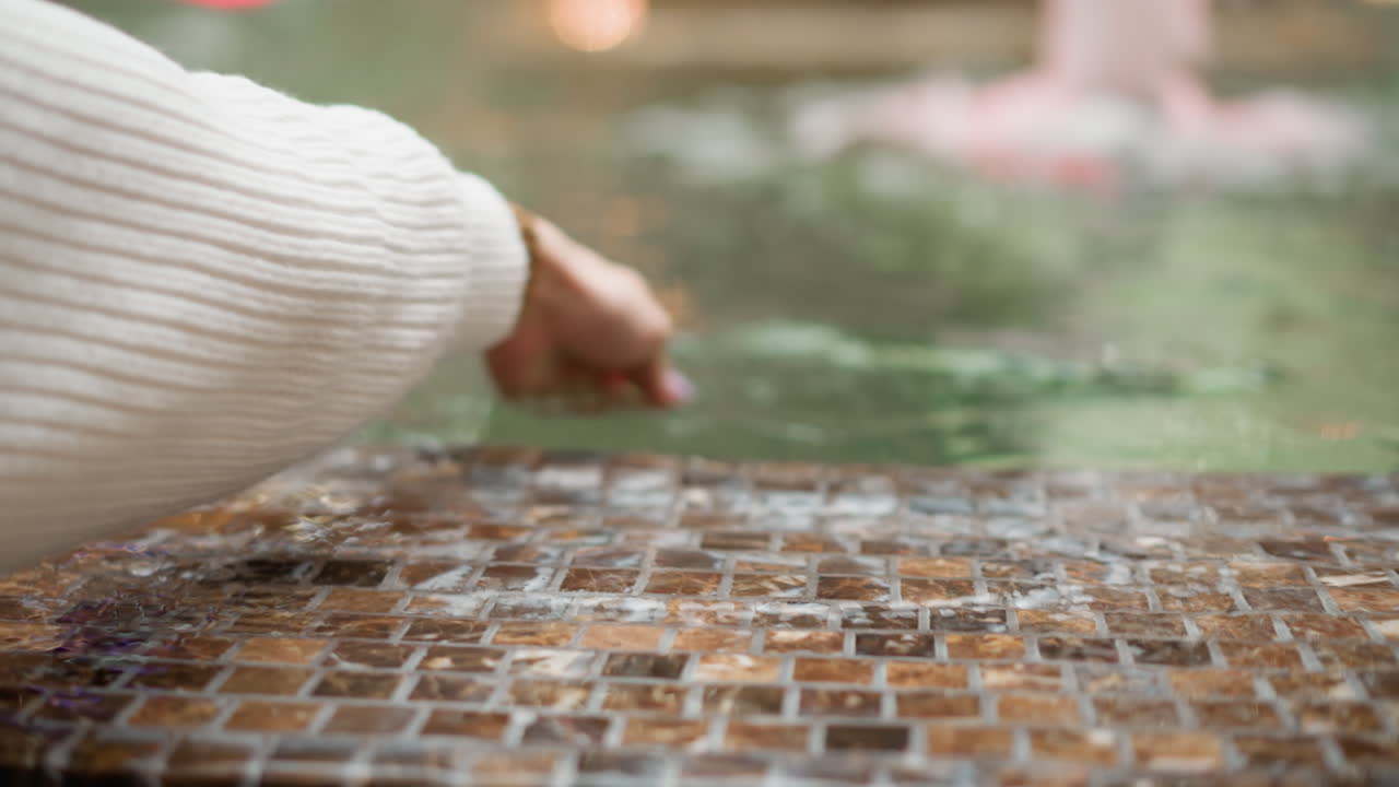 close up of female hands touching water surface in tiled mall fountain under soft interior lighting with gentle ripples and blurred background jets creating subtle serene leisure moment
