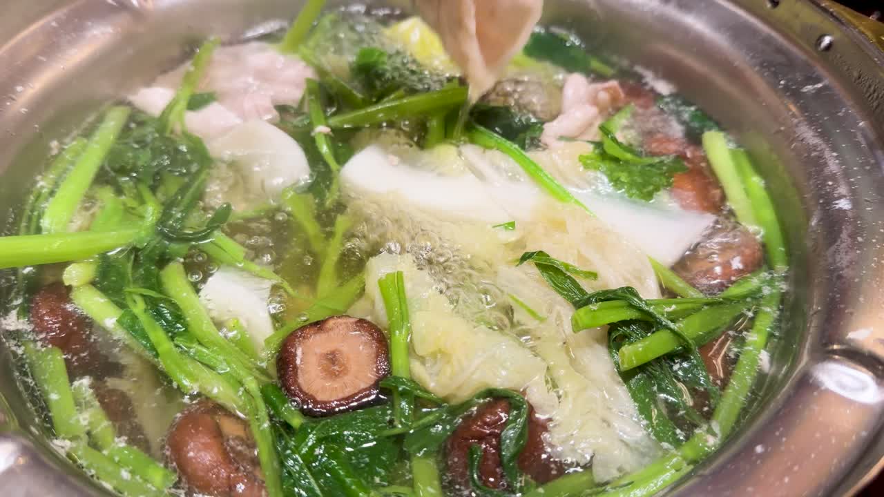 Chopsticks lift cooked meat from steaming hotpot filled with leafy greens, mushrooms, and broth