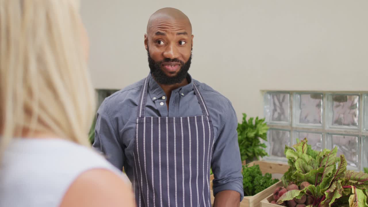 Vidéo d'un vendeur afro-américain heureux montrant des légumes biologiques à une cliente caucasienne.