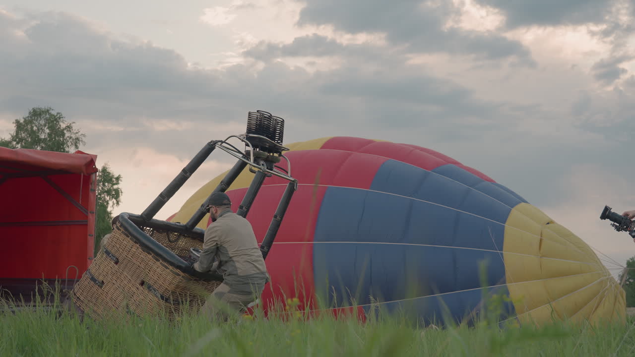 woman steps out from basket frame as man lowers burner assembly gradually against backdrop of deflating balloon envelope at dusk field crew preparing for flight and secure rope lines landing