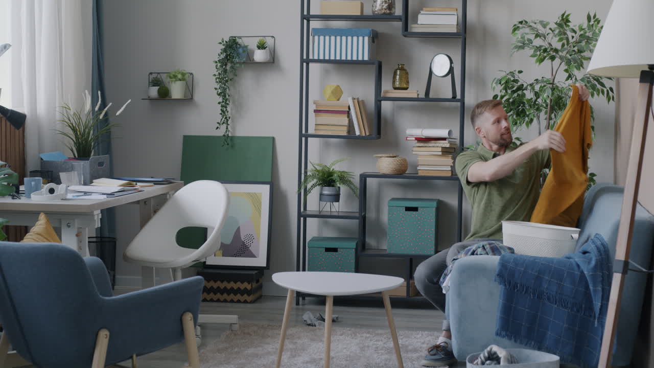 Man Putting Away Clothes on a Couch in a Messy Living Room