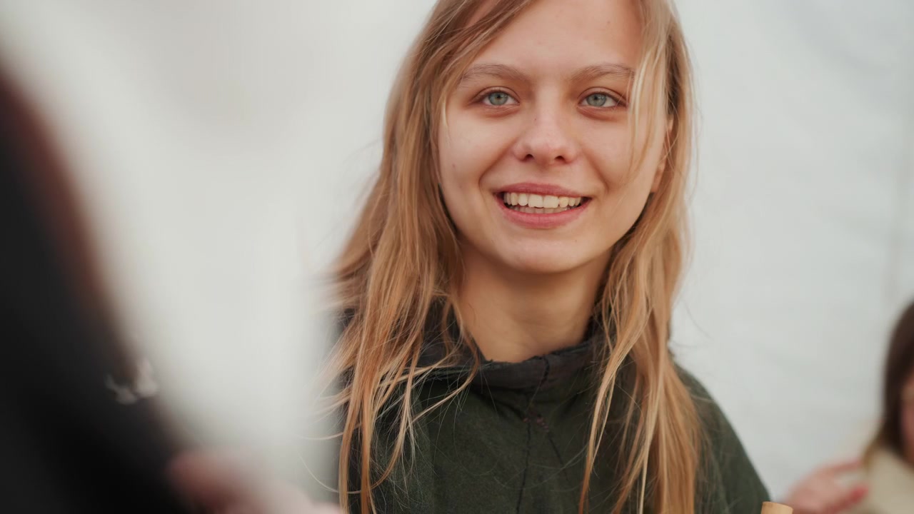 Una giovane donna con lunghi capelli biondi si gira verso lo spettatore e sorride in un ambiente interno illuminato delicatamente, con un leggero movimento dei capelli, una postura rilassata, un contatto visivo gentile e un'atmosfera calda e informale.