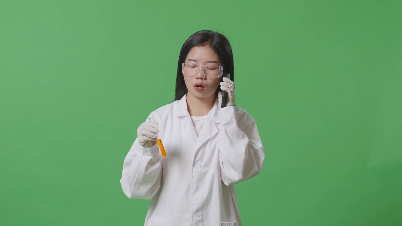 아시아 여성 과학자가 실험실에서 녹색 화면 배경에 서서 스마트폰으로 대화하는 동안 손에 실험관에 있는 오렌지색 액체를 보고 있습니다.