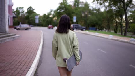 una morena feliz con un suéter verde camina con un patineta en el parque. vista de atrás. caminar en el parque, retrato. pasatiempo, ocio