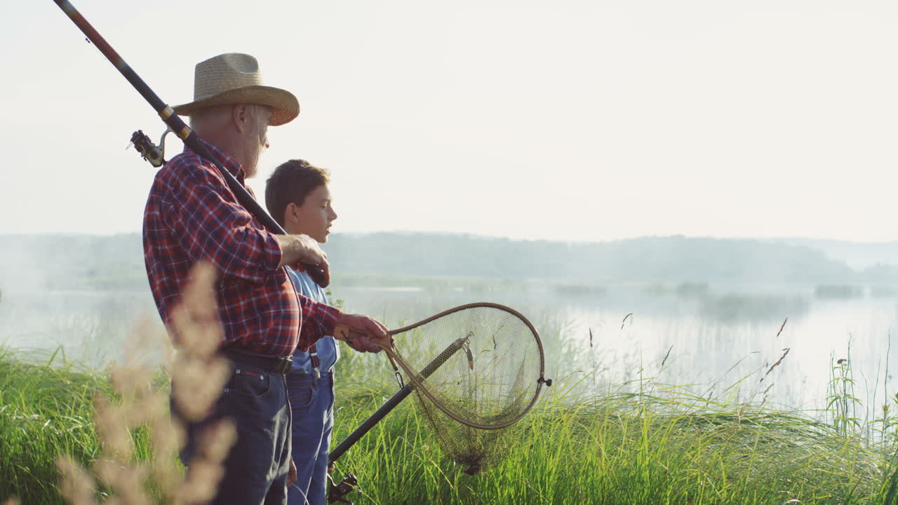 Little teen boy with his grandpa walking on the lake shore with rods and fishing equipment and talking early in the morning