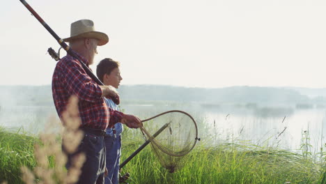 Little-teen-boy-with-his-grandpa-walking-on-the-lake-shore-with-rods-and-fishing-equipment-and-talking-early-in-the-morning