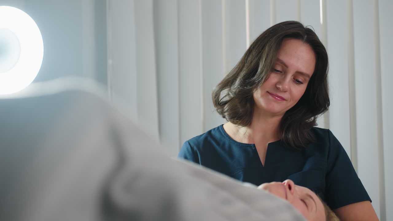 Skin therapist smiles while massaging female client lying on spa bed in bright room with white blinds background during relaxing facial therapy session showcasing expert care and joyful atmosphere