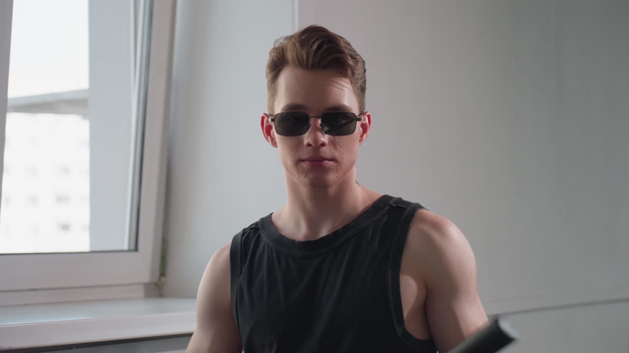 Boy in black tank top and dark shades playfully taps security baton on forearm by loft window, smiling with confident flair, tattooed neck and muscular build