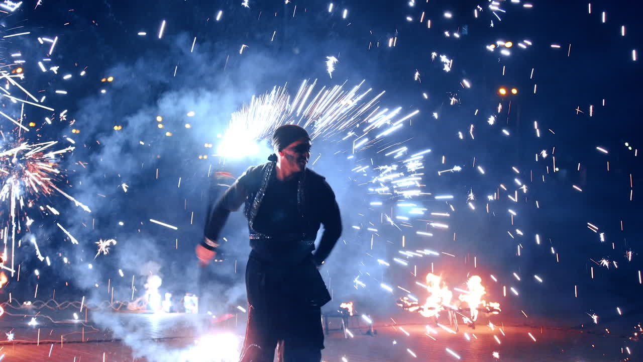 Man in black costume and bandana dances with a stick producing fireworks to the sides. Spectacular fire show at night.
