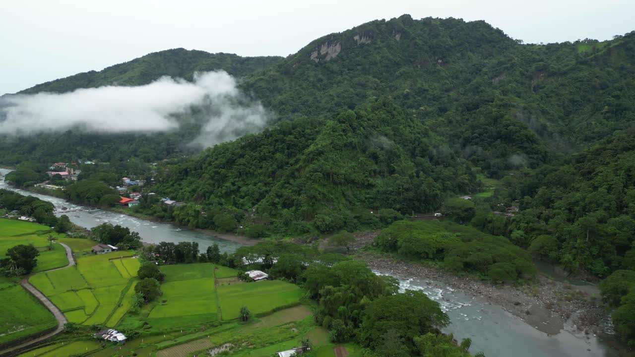 Aerial view of a Philippine valley with vibrant rice paddies, scattered homes, and a winding river framed by misty forested mountains in a peaceful tropical landscape