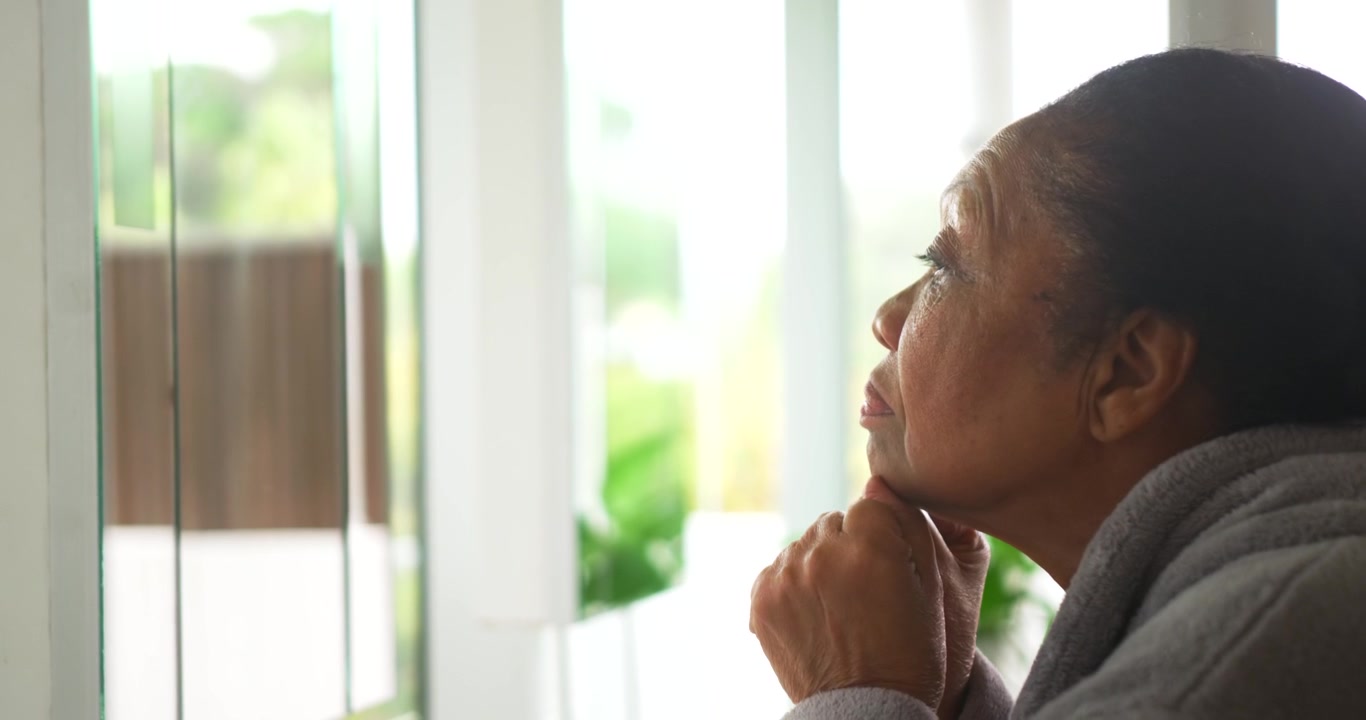 Senior African American woman sitting at blinds at home wearing gray robe seeing yard resting chin