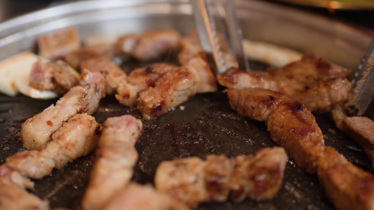 Delle pinze in metallo girano e dispongono fette di pancetta di maiale condita su una griglia rotonda da tavolo.