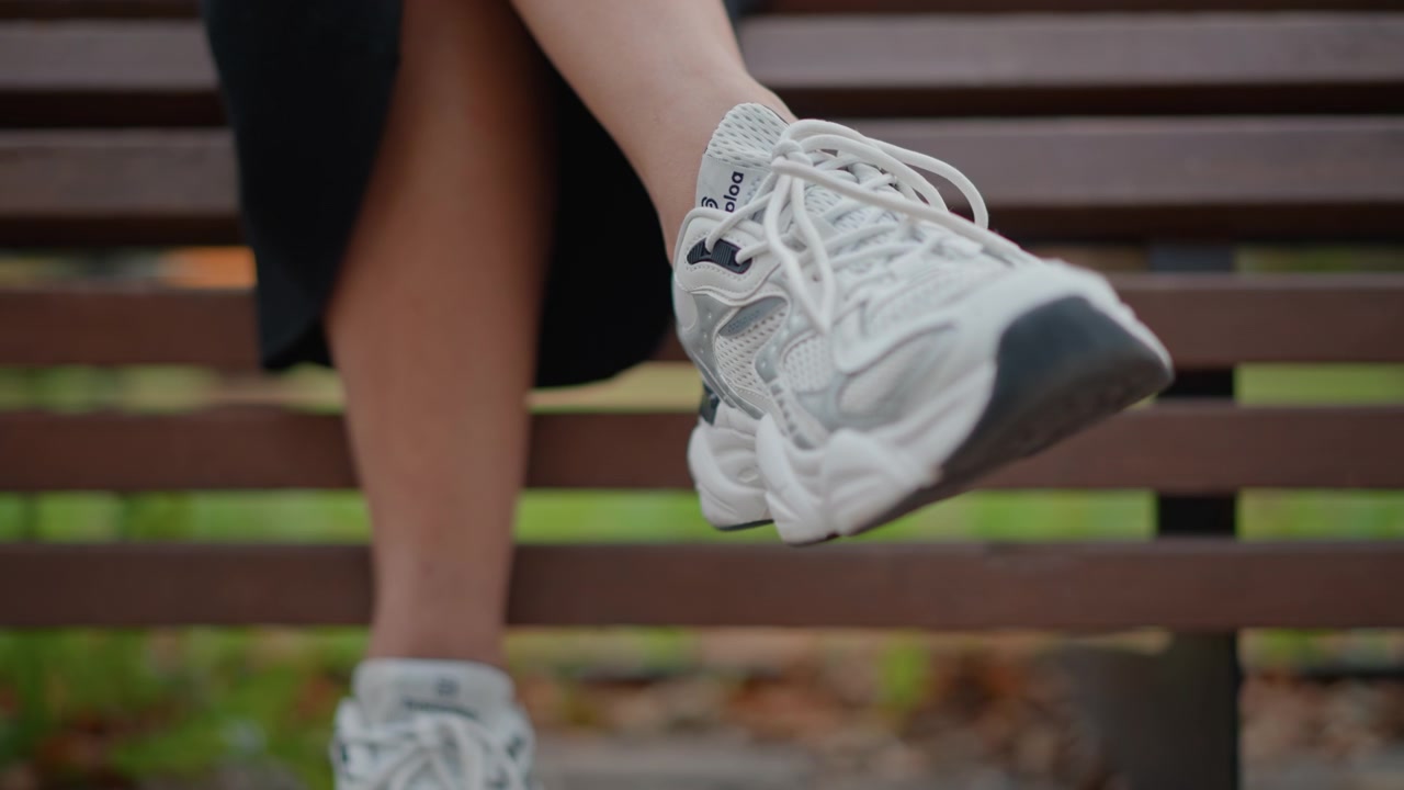 Vista dal basso di gambe incrociate che indossano sneakers bianche massicce, con un piede oscillato in avanti sopra una panchina di legno del parco e la suola chiaramente visibile, creando un movimento seduto rilassato con luce diurna soffusa e
