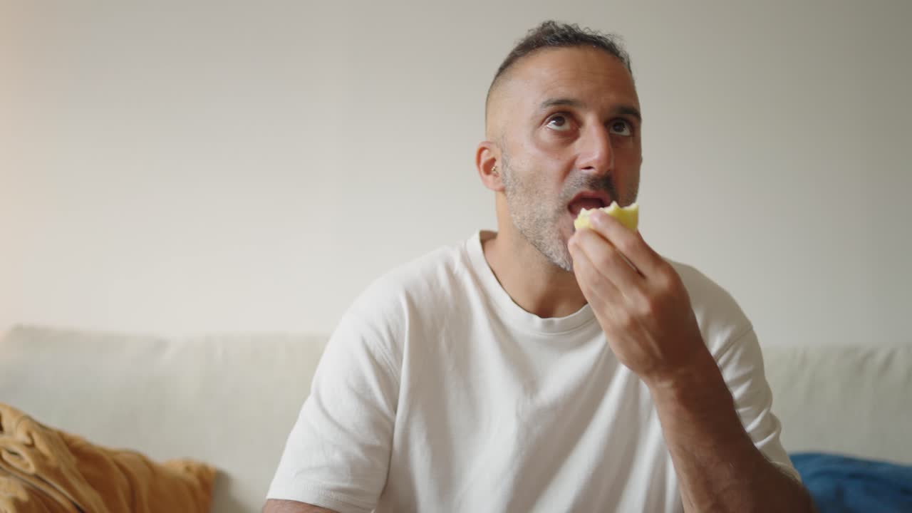 A middle aged man sits facing the camera, eating fruit while watching television, capturing the mood of ordinary and monotonous daily life