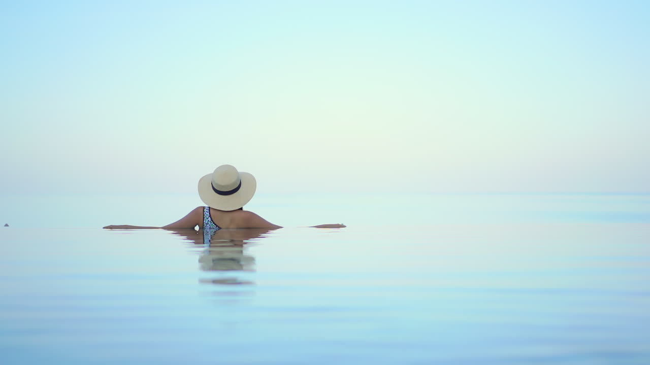 Helle, ätherische Aufnahme einer Frau von hinten, die sich in einem Infinity-Pool entspannt.