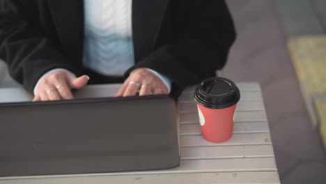 close-up of hands typing on laptop keyboard on outdoor table, person wearing black coat, white sweater, and rings while coffee cup is next to laptop