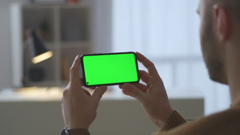 pantalla verde en un teléfono inteligente moderno en manos de un hombre sentado en casa y viendo video en primer plano a través del hombro del usuario masculino