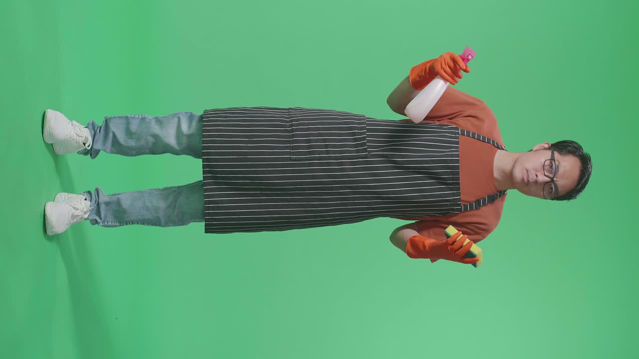 아시안 남성 집주인이 스프레이와 주방 스폰지를 손에 들고 초록색 스크린 스튜디오에서 머리를 흔들고 있습니다.