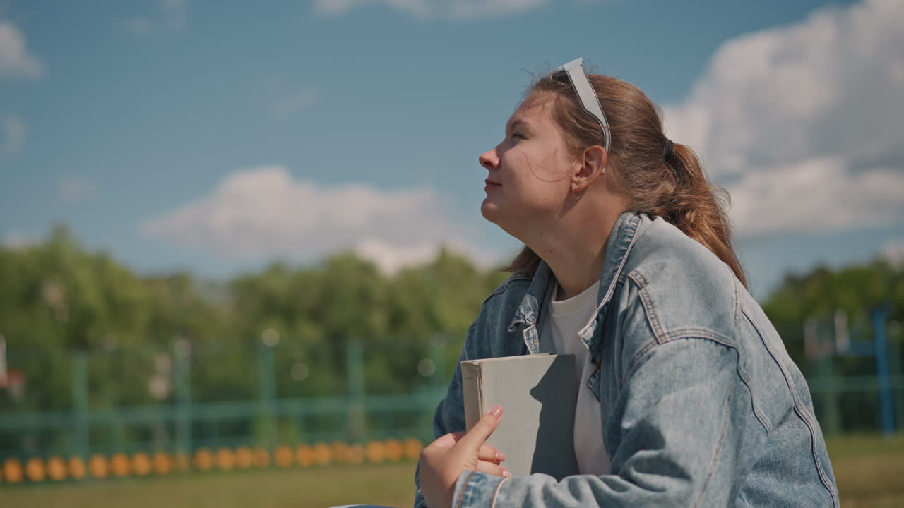Joven relajada en un paisaje de parque, sentada en un campo de hierba, estudiando tranquilamente bajo la luz del sol. Viste chaqueta de mezclilla y mira al cielo durante una sesión de estudio al aire libre soleada.