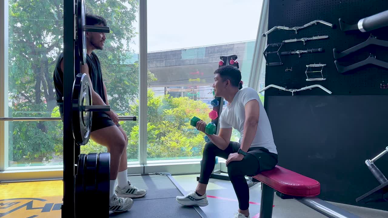 Pelatih mengawasi klien yang melakukan seated dumbbell curls di gym modern yang terang dengan jendela besar