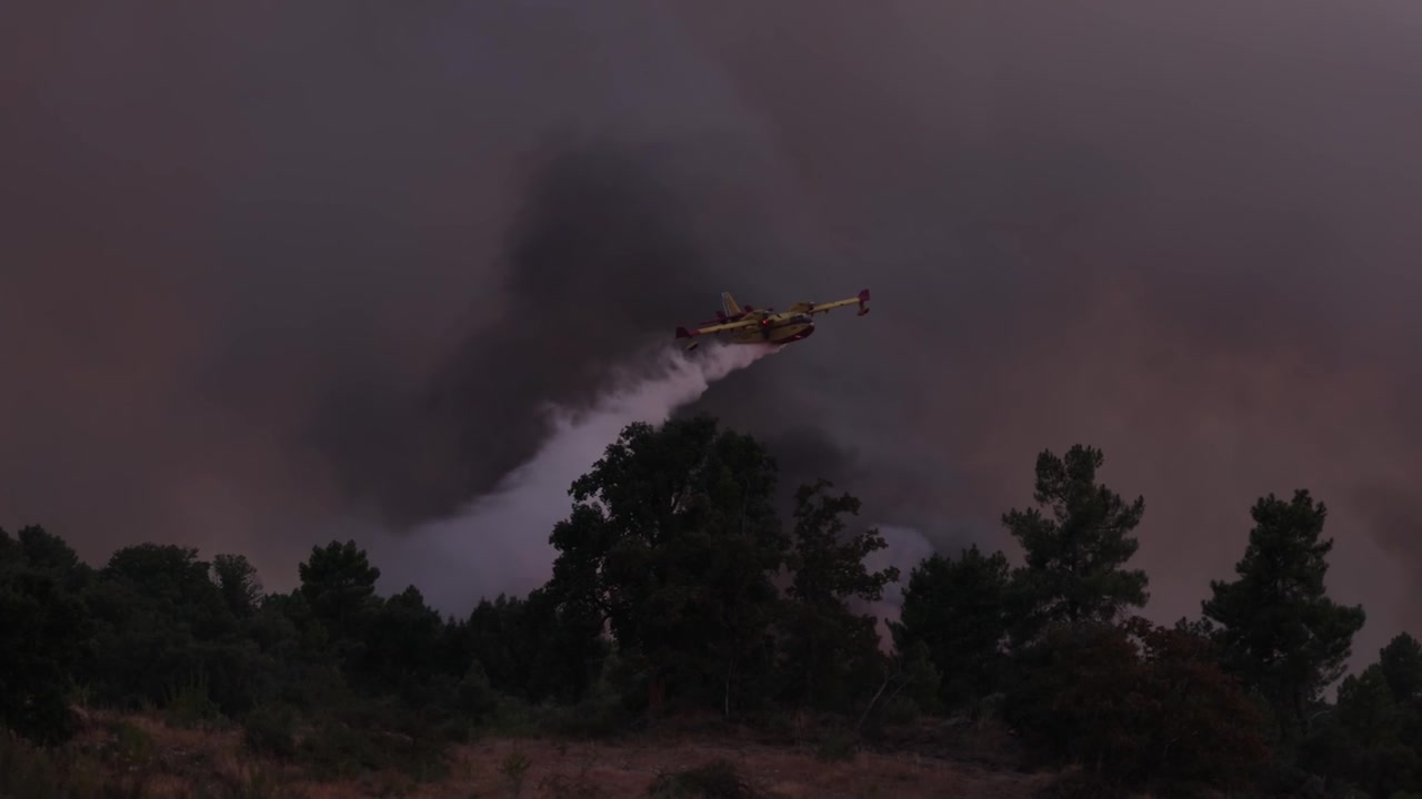Espectacular toma de un avión contraincendios Canadair CL-215T descargando agua sobre un incendio forestal en España, con espeso humo elevándose bajo los sombríos cielos de verano del fenómeno de El Niño