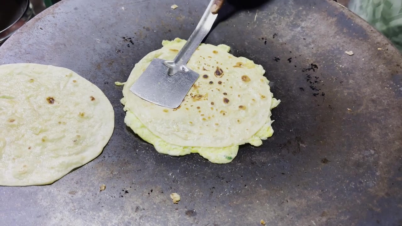 Omelette fresca che viene girata su una padella calda accanto a del pane piatto, con un'ambientazione da cucina rustica, una scena di preparazione di cibo da strada e un'atmosfera di cucina tradizionale!
