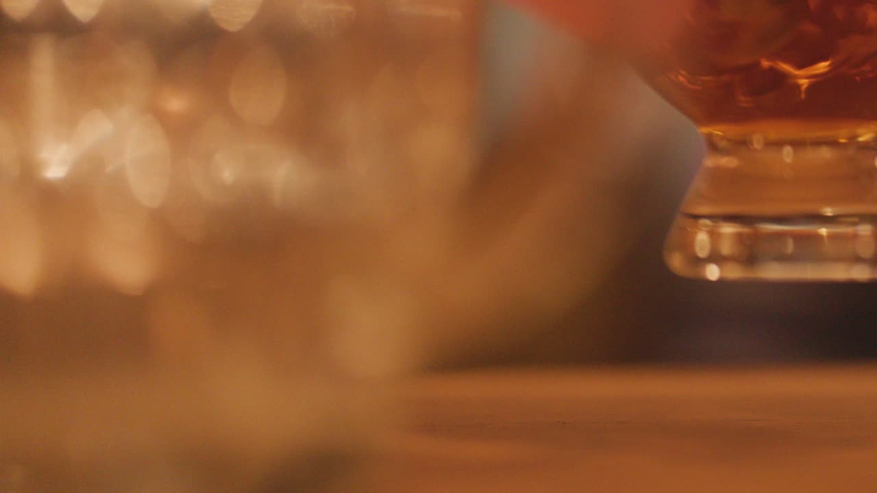 A hand reaches for a whisky glass on a wooden table, surrounded by blurred candlelight, creating a warm, intimate nighttime atmosphere with shallow focus