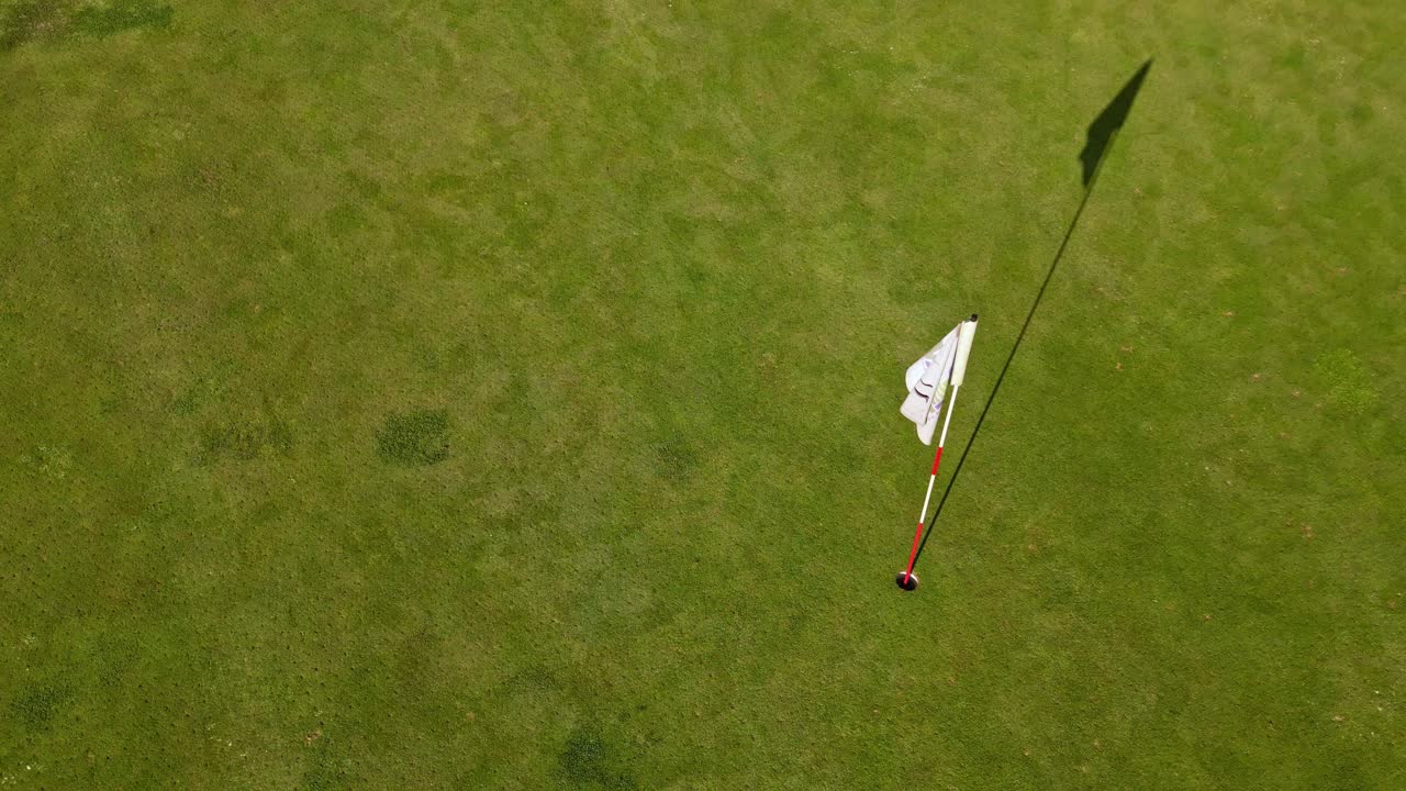 Descending camera over well-maintained golf course green, revealing a white flagstick casting long shadow across the smooth grass, begining elevated and gradually lowers toward the putting surface