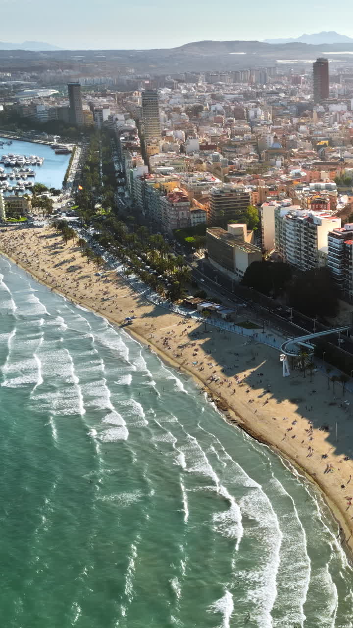 Luftdronebillede af biler, der kører langs kystlinjen, med folk der slapper af på stranden i Alicante, Spanien i dagslys. Lodret