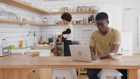 busy family in kitchen at breakfast with father working on laptop and mother caring for baby son