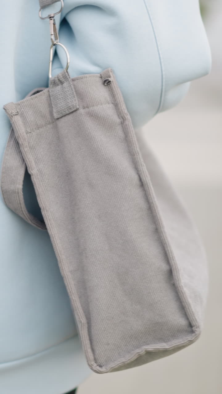 Back view of young girl in casual blue hoodie holding gray bag while walking majestically outdoors with blurred background of iron fence and green grass