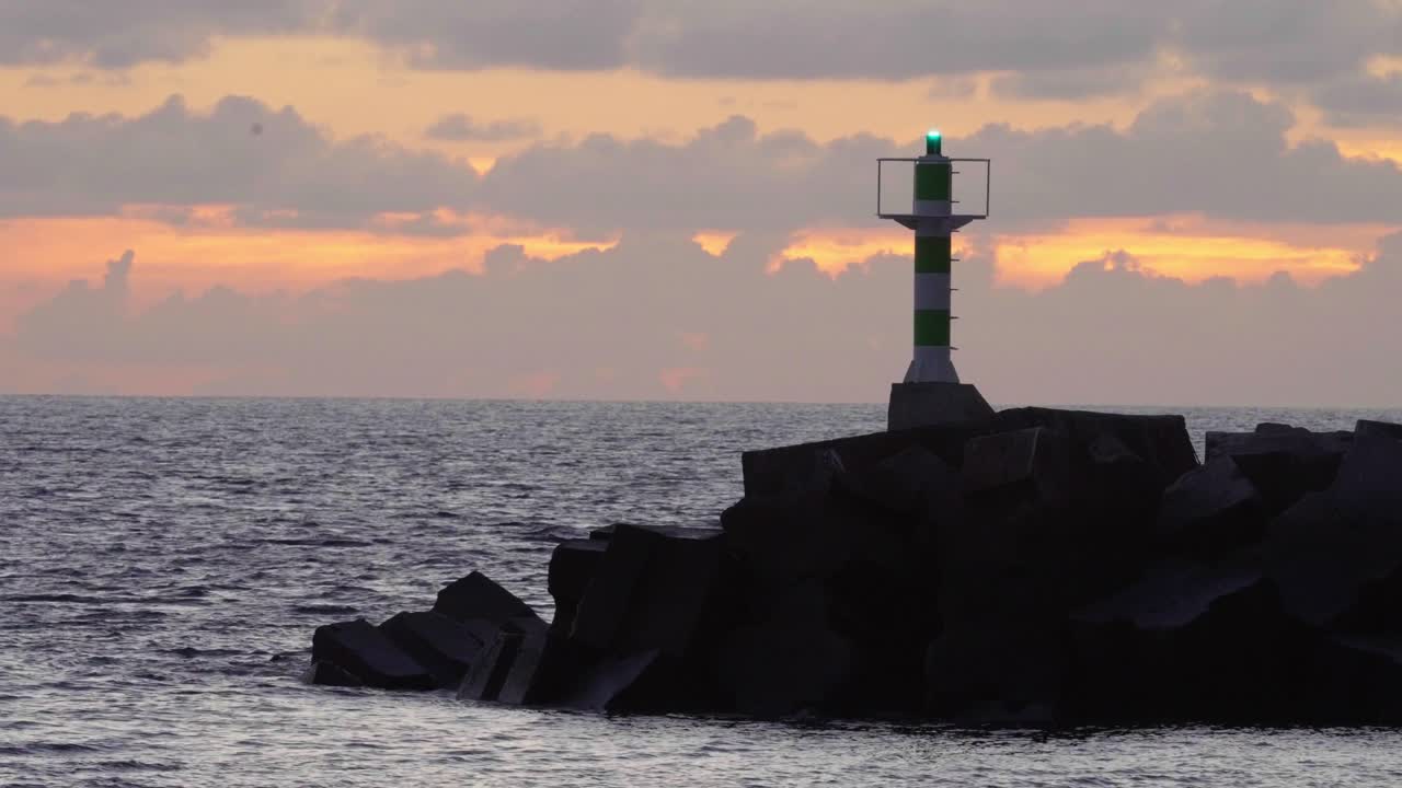 A lighthouse stands tall on rocky cliffs as the sun sets behind it, with waves crashing in fast motion. The golden glow of the sky transitions quickly, creating a dynamic and serene coastal scene.