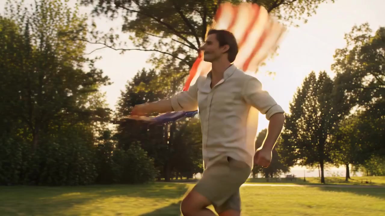 Running man waving large American flag in sunlit park as breeze makes flag billow, golden light