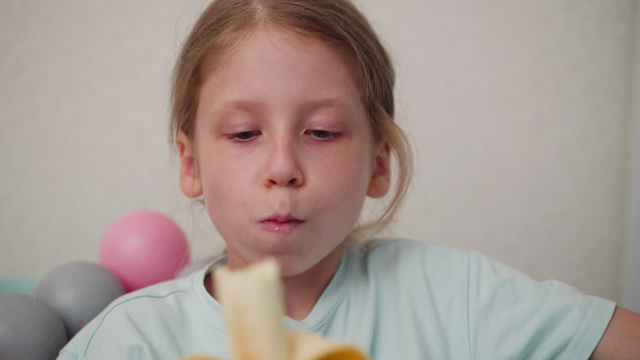 Close up of little girl savoring banana while sitting in pastel ball pit, holding peeled fruit gently with both hands, focused expression captured with soft lighting