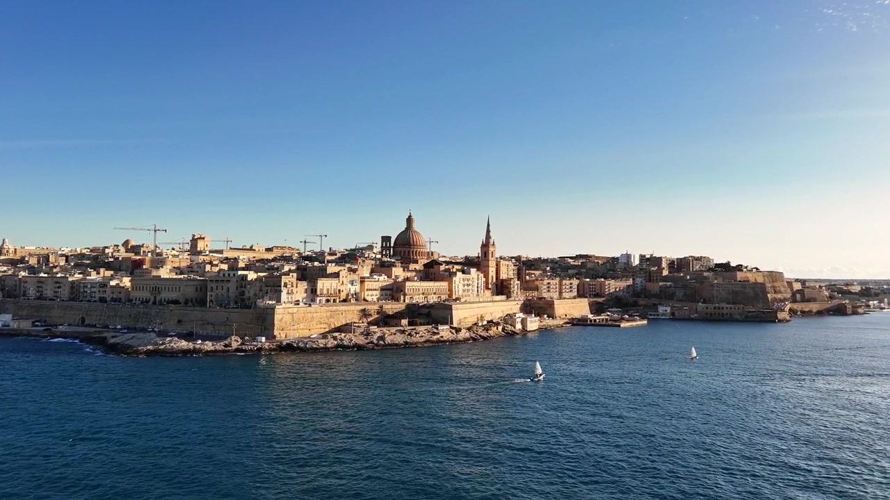 La Basilica di Nostra Signora del Carmelo si erge sullo skyline di La Valletta, Malta, con edifici in pietra calcarea e il lungomare del Grand Harbour, mentre la telecamera si avvicina lentamente, con un drone che avanza dal Mar Mediterraneo