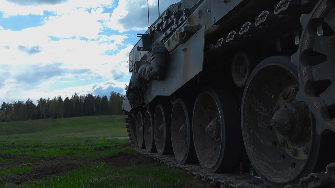 Low angle close up or closeup footage of a British army or military Challenger 2 tank tracks while the armored vehicle is standing on a grassy green field and aiming its cannon towards a treeline.