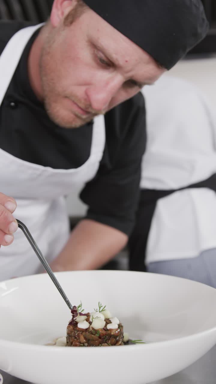 Focused caucasian male chef decorating prepared meal on plate in kitchen, slow motion, vertical