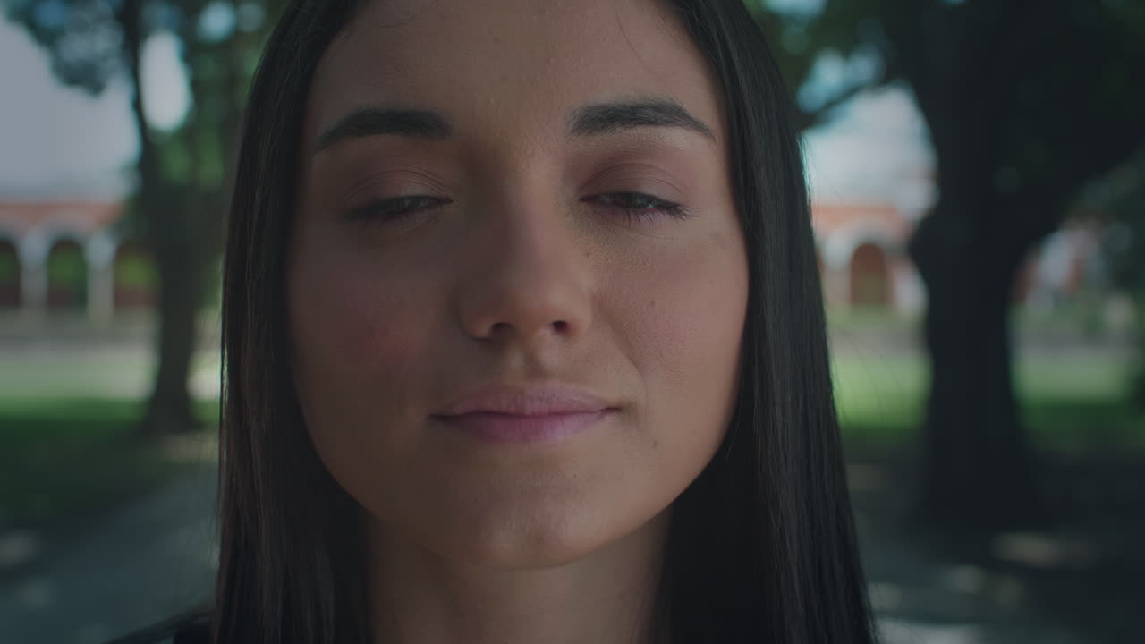 High-quality 4K shot of a young brunette in natural light, slowly opening her eyes in a park setting. She has a peaceful, daydreaming expression. Ideal for nature, mindfulness,