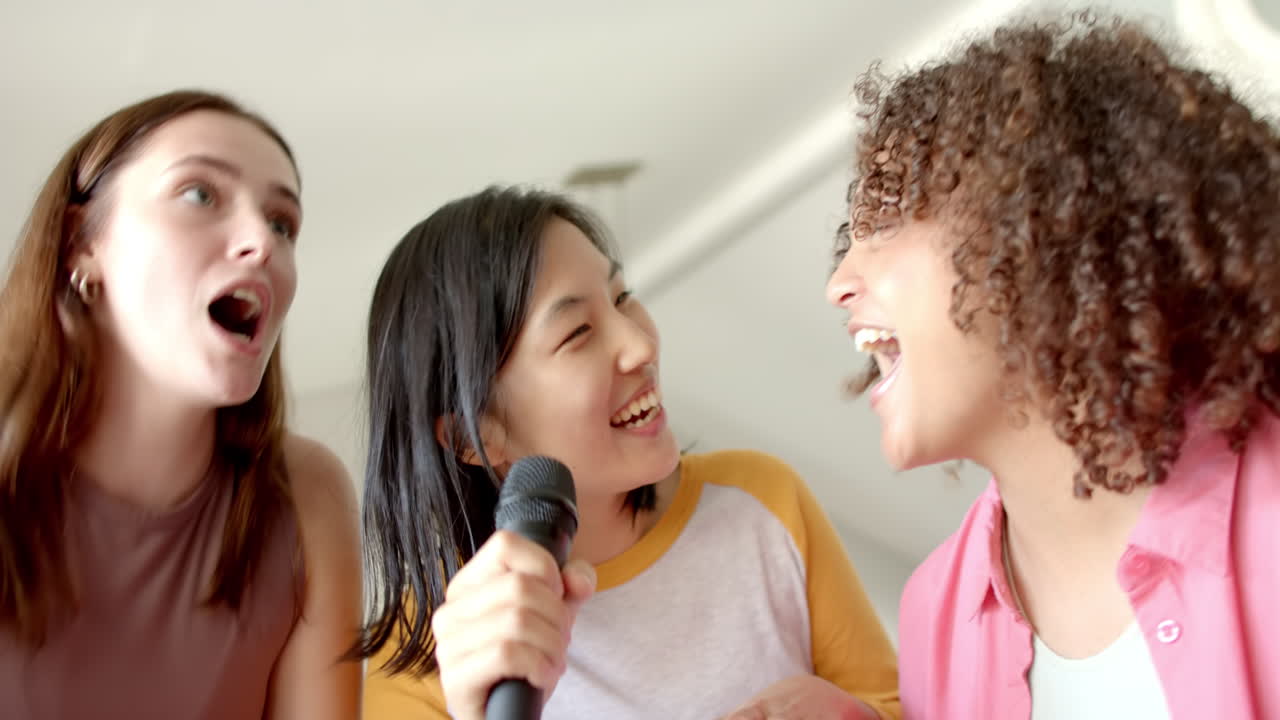Singing together, three women friends enjoying karaoke at home, holding microphone