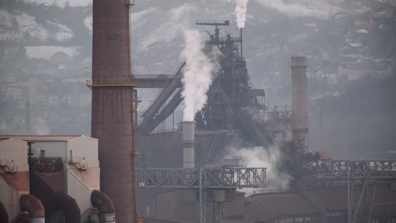 Industrial smokestacks emit plumes of smoke, with a backdrop of a snowy hillside and urban landscape