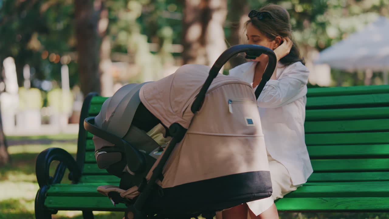 Young mother sitting on a park bench next to a stroller, enjoying a peaceful outdoor moment with her baby