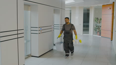 arabic cleaning man holding cleaning products and mop bucket while walking down a corridor inside an office building