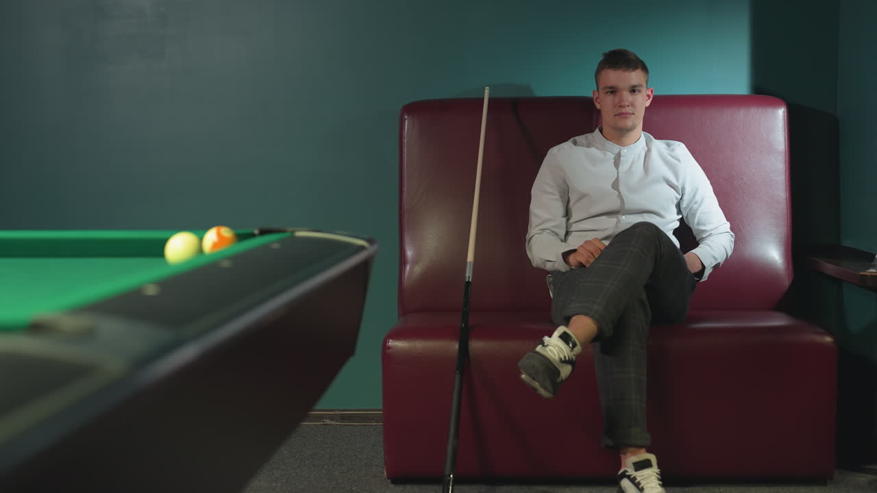 un hombre con camisa blanca y pantalones a cuadros se sienta en un sofá rojo, rodando una pelota de billar naranja en la mano con una expresión seria.