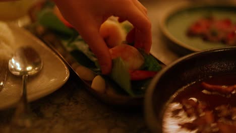 a hand assembles miang kham with shrimp on a table in warm lighting, highlighting the vibrant ingredients and intricate preparation
