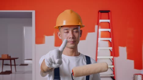 primer plano de un pintor asiático que lleva un casco de seguridad apuntándote y desaprobando sin señal del dedo índice mientras está de pie pintando la pared en la habitación