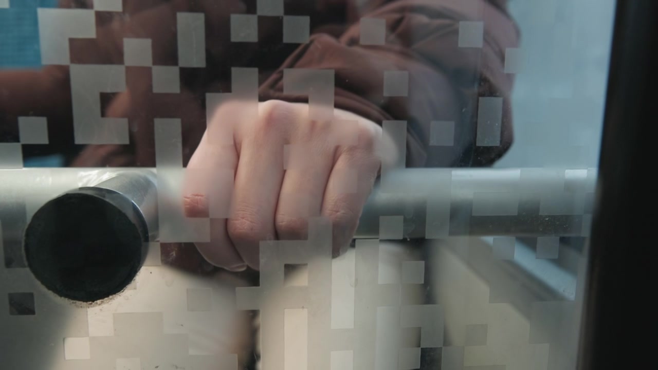 Vista frontal cercana de una mano derecha sujetando firmemente una barandilla horizontal de acero inoxidable detrás de un cristal esmerilado con patrón cuadrado para privacidad, mostrando presión, soporte, textura y reflejos interiores tenues.
