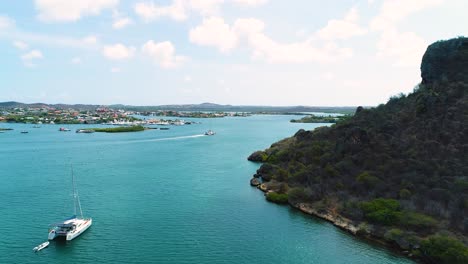 Catamaran-anchored-in-Spanish-Waters-of-Curacao-off-of-dry-coastline-as-boat-drives-in-distance,-aerial-dolly